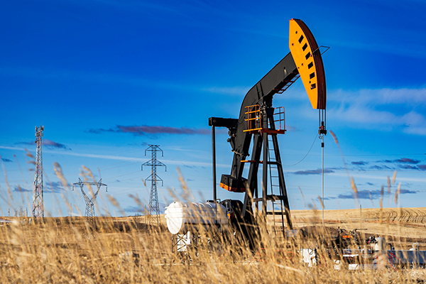 An oil pumpjack operating in a golden prairie field under a blue sky, with power lines and transmission towers in the background.
