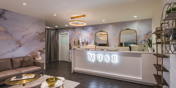A modern and elegant reception area at Muse Aesthetics featuring a marble-patterned accent wall, a white front desk with a backlit sign, gold mirrors, and seating with decorative elements, creating a warm and luxurious atmosphere.
