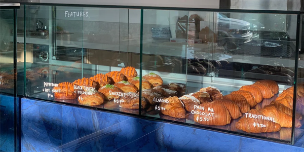 A glass display case filled with a variety of croissants at Otie, including traditional, pain au chocolat, almond, Snickers, black truffle pecorino, and pepperoni hot honey, with handwritten labels on the glass.